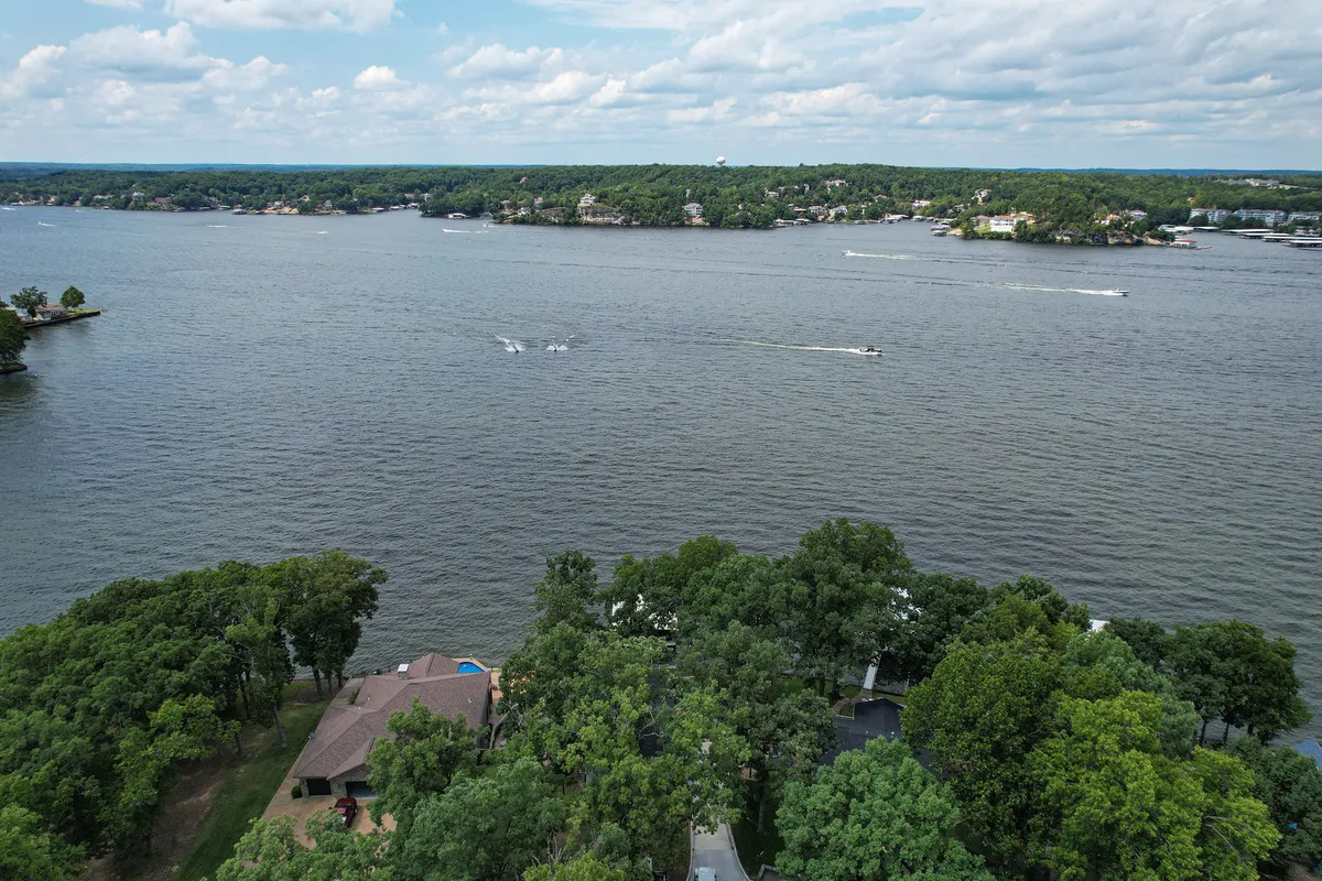 This scenic view showcases the lake, framed by trees, with houses and boats visible. The tranquil setting highlights the lake's activity, with boats gliding by and abundant nature all around, capturing the energy and beauty of Lake of the Ozarks.