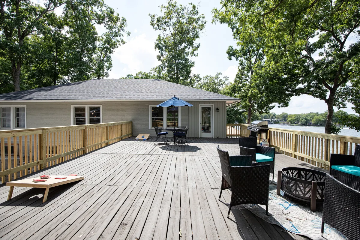 The image presents a wooden deck furnished with outdoor seating, an umbrella, a grill, and a panoramic view of the water. The environment is cheerful and primed for socializing, alfresco dining, and lakeside sunsets.