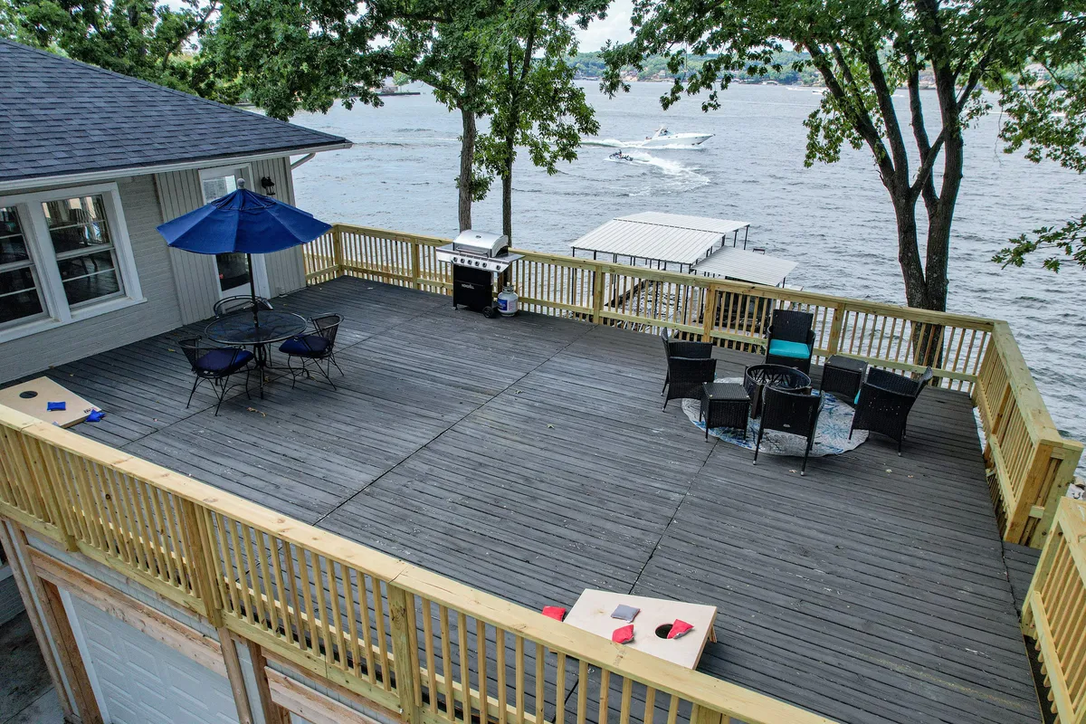An exterior deck area overlooking the lake, featuring outdoor seating, a grill, and lush trees. The spacious deck is staged for both relaxation and social activity, offering an ideal spot for barbecues, dining, or playing games against a scenic lake backdrop.