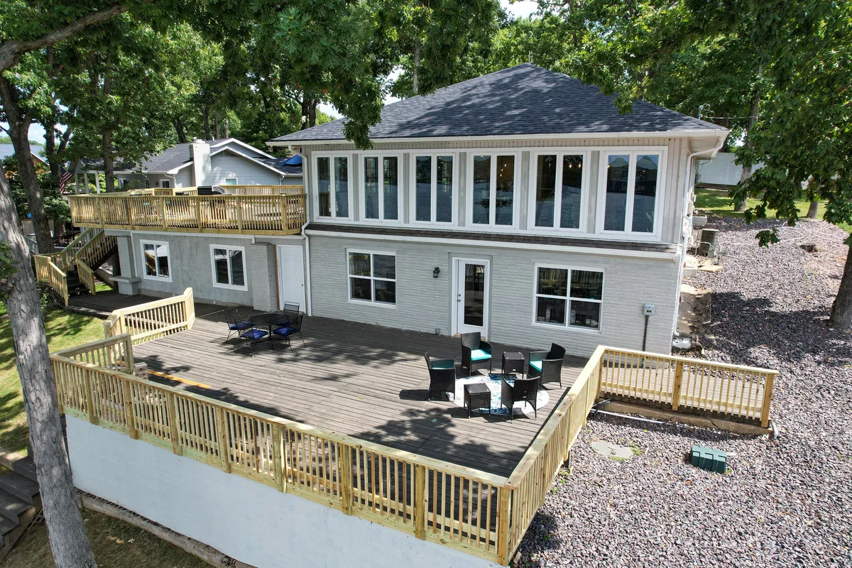 A large deck with ample outdoor seating, overlooking a landscape of trees and partially visible water. The space is designed for family gatherings, relaxation, and meals, set against a natural backdrop ideal for both morning coffee and evening barbecues.