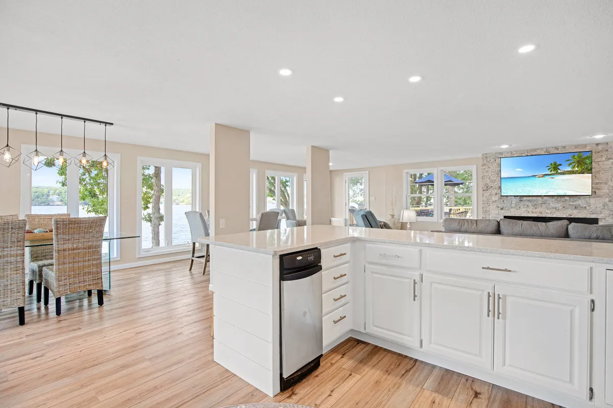 A bright, open-plan kitchen and dining area featuring a large island with white cabinets, woven dining chairs, and expansive windows offering beautiful lake views. The space also includes a TV mounted on the wall and a comfortable living area, creating a seamless environment for cooking, dining, and relaxing.