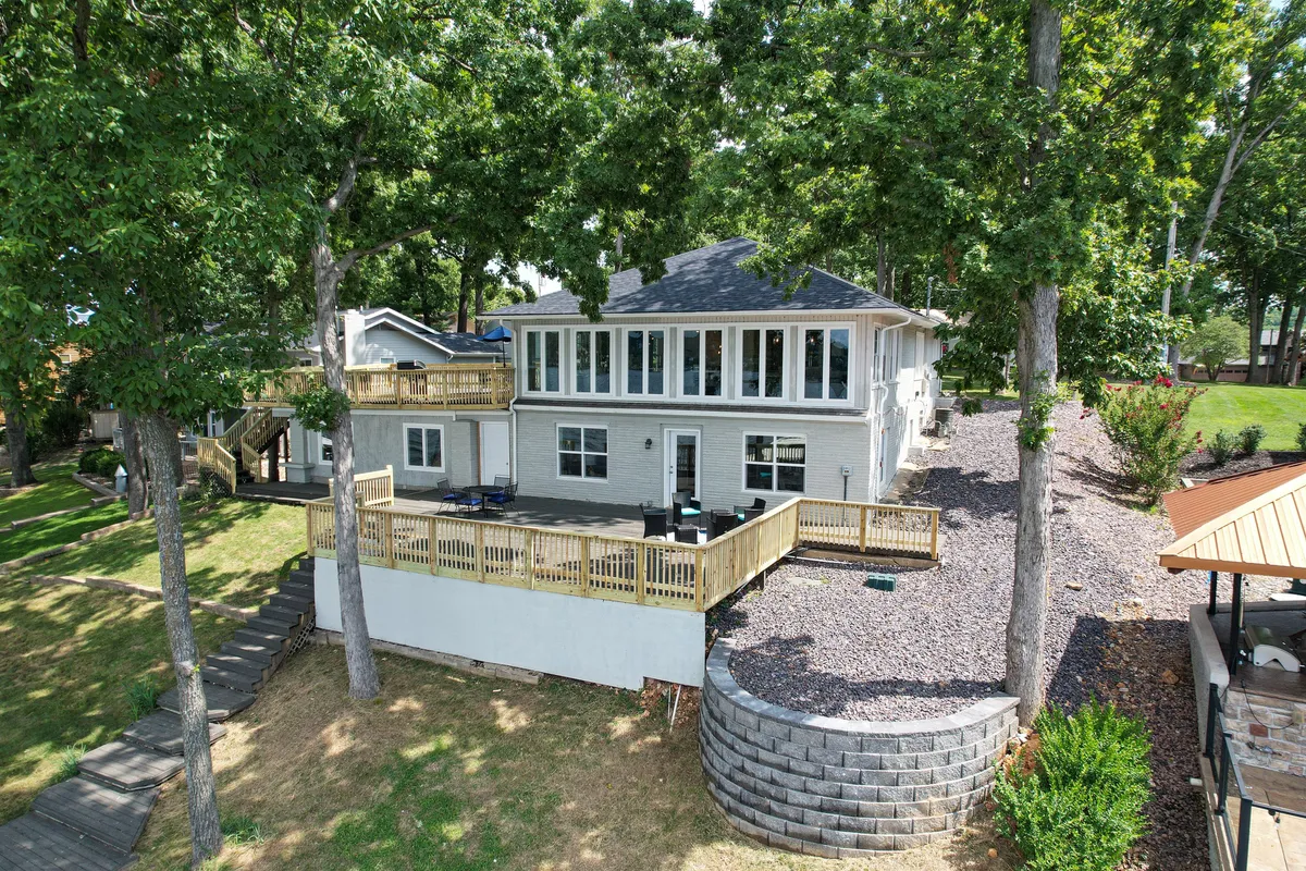The image features a spacious single-family lake house framed by lush trees, with two expansive decks outfitted for outdoor dining and relaxation. The decks overlook the sparkling waters of Lake of the Ozarks, with abundant seating inviting guests to unwind, soak in sunset views, and enjoy al fresco meals or lively gatherings with friends and family. Large windows on the home promise natural light and picturesque lake vistas from inside.