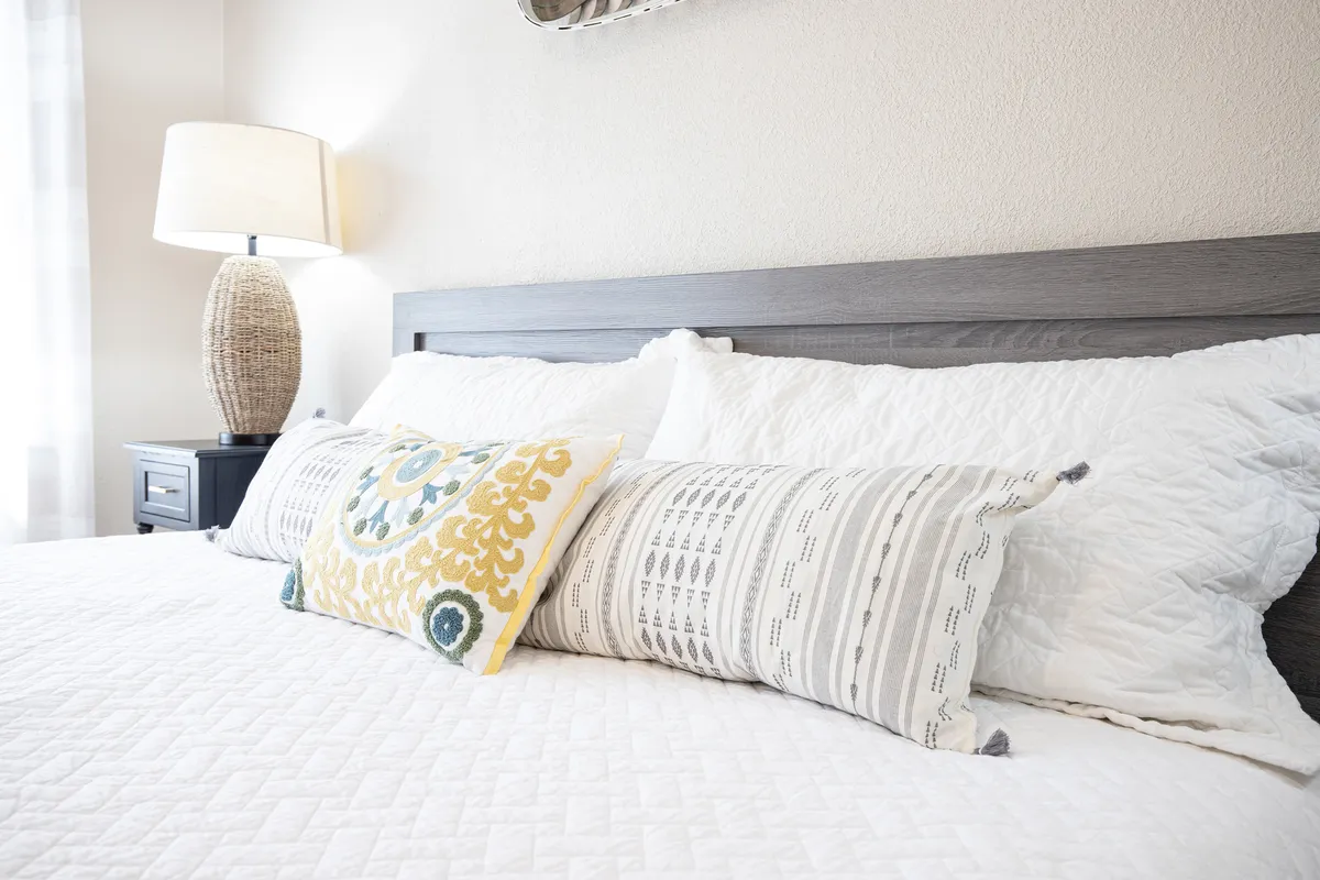 A cozy bedroom featuring a neatly made bed with colorful decorative pillows, a woven lamp, and a welcoming atmosphere. Soft lighting and thoughtful design create a serene space ideal for restful nights or a quiet reading session.