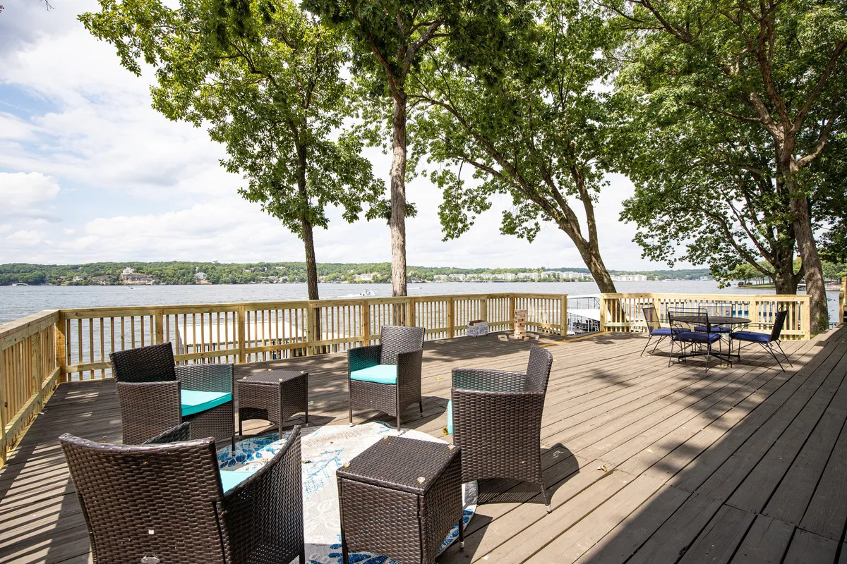 This image captures an inviting outdoor seating area overlooking the water, with comfortable furnishings and natural shade from surrounding trees. The deck offers a tranquil setting, perfect for relaxing, enjoying meals outside, or spending time with loved ones while soaking up the lake view.
