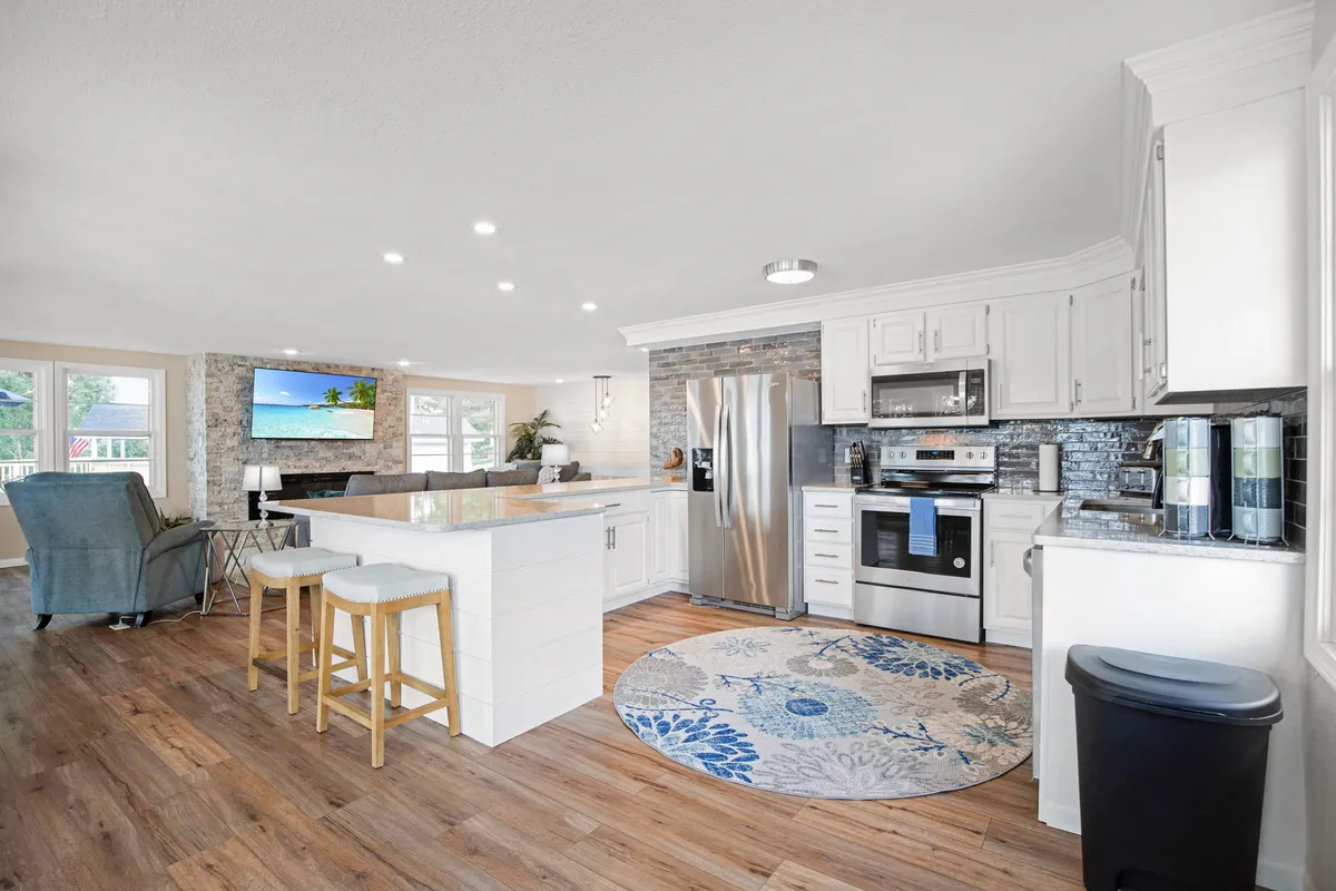 A cozy kitchen with an island, visible appliances, a decorative rug, and a television. The setup is casual and welcoming, supporting both cooking and relaxed family gatherings. The kitchen is open to an adjoining living and dining area.