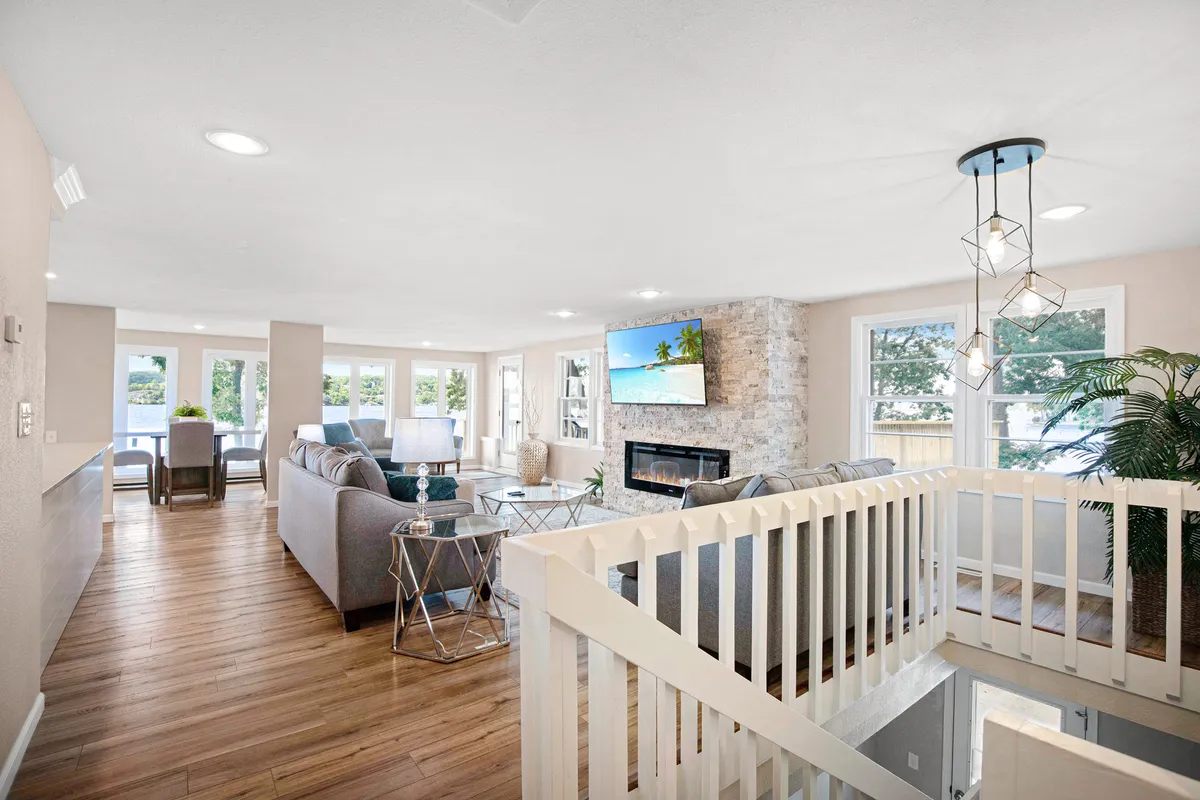 A spacious, modern living room with a stone fireplace, large windows framing the lake, and comfortable, contemporary furniture. The room exudes warmth, with ample seating ideal for group gatherings and sunny afternoons spent lounging. A TV set above the fireplace adds to the possibilities for entertainment and relaxation.