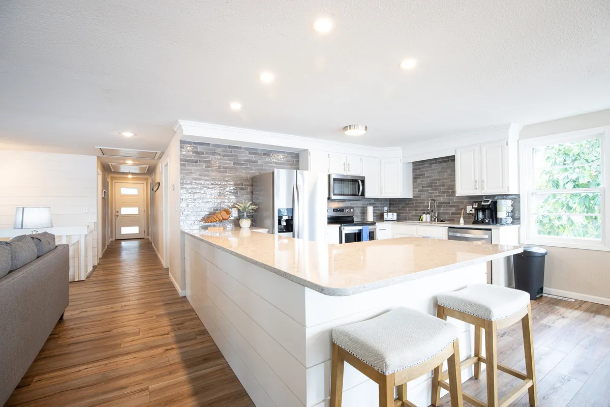 A modern kitchen featuring a spacious island counter, stainless steel appliances, a sleek grey stone backsplash, and bar stools. The space looks inviting and is clearly designed for both cooking and socializing, echoing the idea of a fully equipped, open kitchen for gatherings.