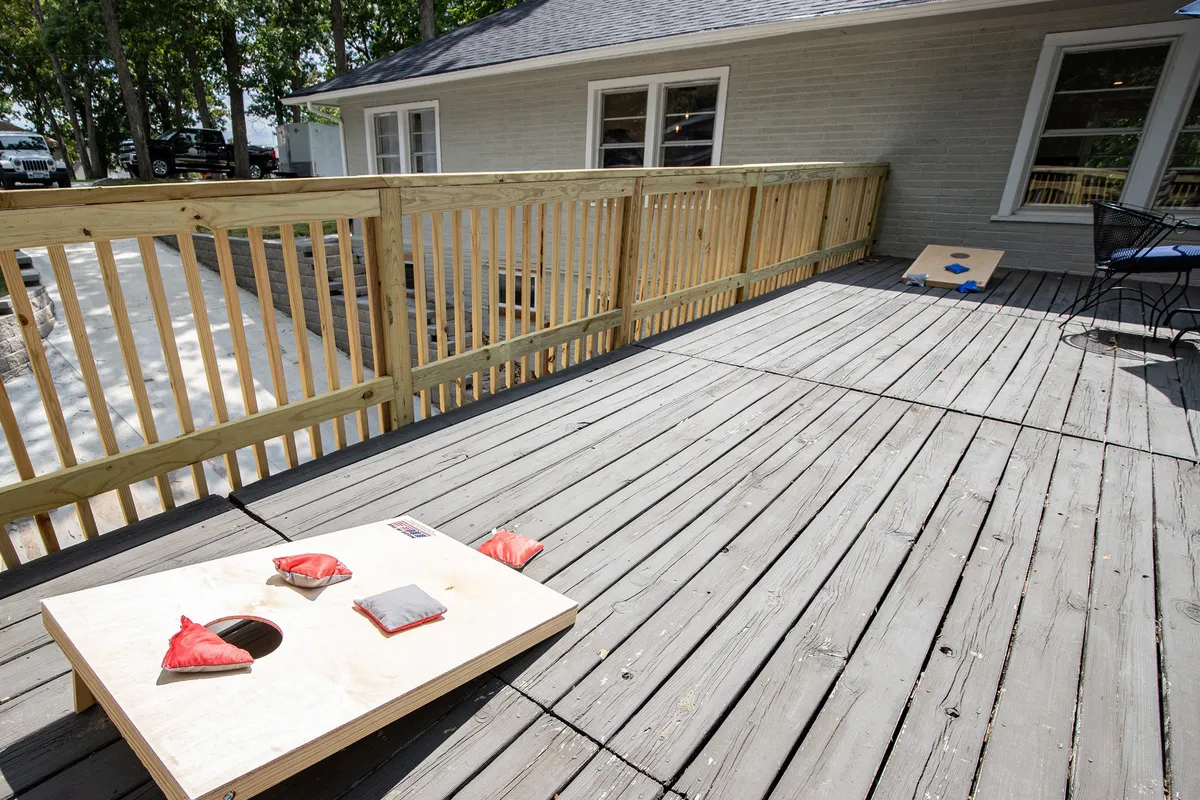 A playful exterior deck scene with a cornhole game set up and surrounding trees. Relaxing chairs suggest a laid-back atmosphere for friendly competitions or casual gatherings as the sun sets over the lake.