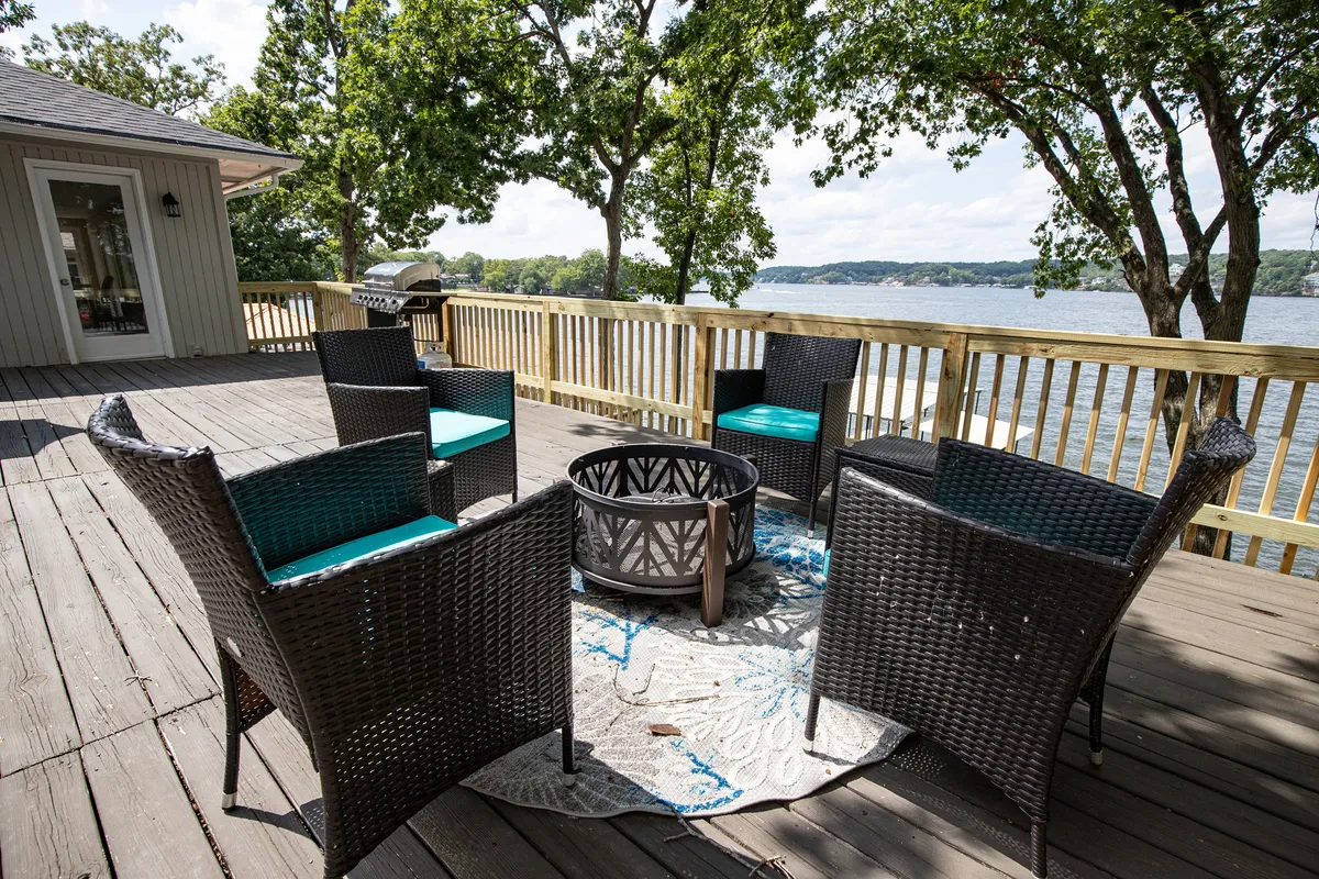 A lakeside seating area with wicker chairs, a fire pit, a patterned area rug, and water views. The scene is set for evening relaxation, socializing, or stargazing, emphasizing comfort and shared experiences by the fire.