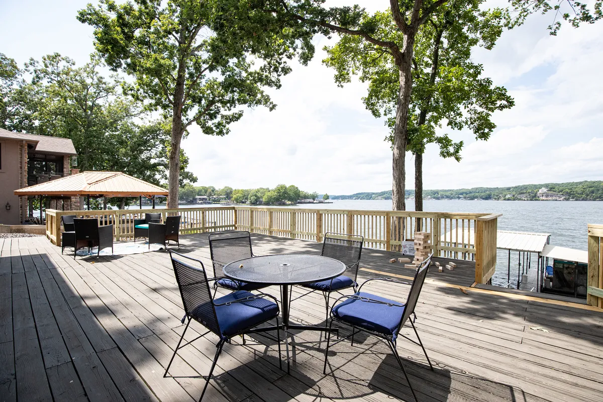 This exterior image captures a table with chairs set on a deck overlooking the water, framed by surrounding trees. The scene is inviting, with the serene lake stretching out in the background, suggesting a perfect space for outdoor dining, relaxing, or watching the sunset.