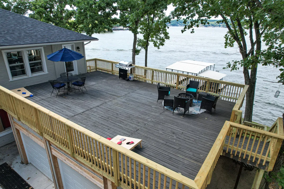 A spacious deck overlooking the water, featuring a comfortable seating area with an umbrella, a grill, and a tranquil lake view in the background. The setup invites outdoor dining, socializing, and enjoying sunsets by the water.