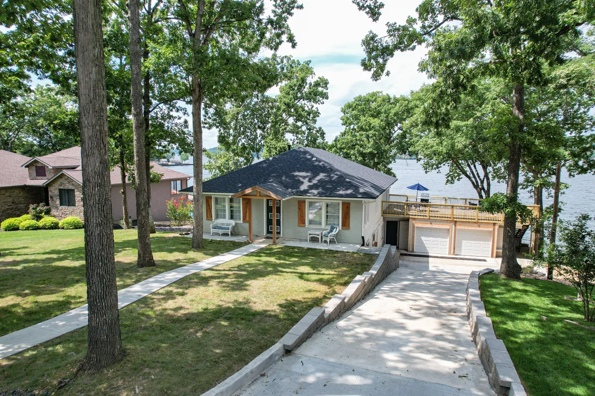 This exterior image showcases the house with its welcoming porch, spacious driveway, vibrant trees, and a scenic view of the lake beyond. The setting highlights areas for gathering, relaxing outdoors, and easy group comings and goings.