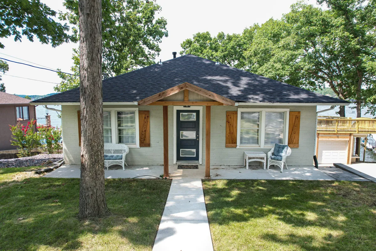 A welcoming exterior shot of the home's front elevation, featuring a charming porch with comfortable seating, manicured landscaping, and a picturesque entryway. Large windows suggest abundant natural light inside, and the porch offers views toward the lake, inviting guests to relax outdoors.