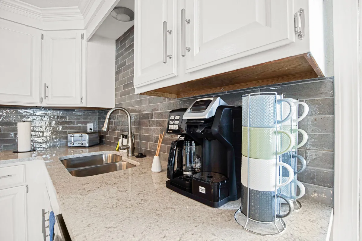 A close-up view of a modern kitchen counter with a coffee maker, neatly stacked mugs, and a sink, suggesting readiness for guests to enjoy fresh coffee and simple amenities.